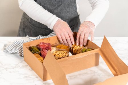 Foto de En el proceso de ensamblaje, una mujer expertamente cura esta caja de charcutería, mostrando carne en rodajas, queso, galletas y uvas, todo arreglado y empaquetado en una caja de regalo marrón. - Imagen libre de derechos