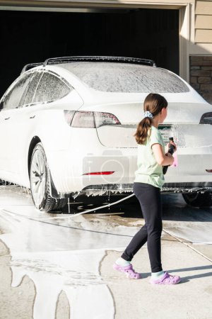 Téléchargez les photos : Une jeune fille aide avec enthousiasme à laver la voiture électrique familiale dans leur allée de banlieue. - en image libre de droit