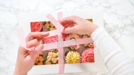 Téléchargez les photos : Pose plate. Dans une main de femme, une boîte en papier blanc remplie de cupcakes gourmands, chacun orné de roses vibrantes et de tulipes fabriquées à partir de glaçage à la crème au beurre, est doucement fermée. - en image libre de droit