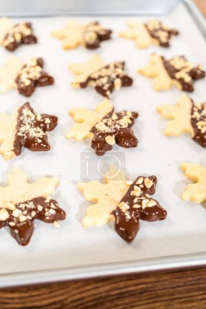 Photo for Creating cutout sugar cookies, partially dipped in chocolate and topped with hazelnut pieces, placed on parchment paper. - Royalty Free Image