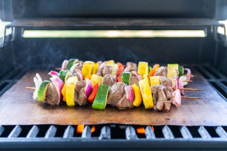 Photo for Skewered beef and fresh veggies sizzle on a copper grill mat over a gas outdoor grill. - Royalty Free Image