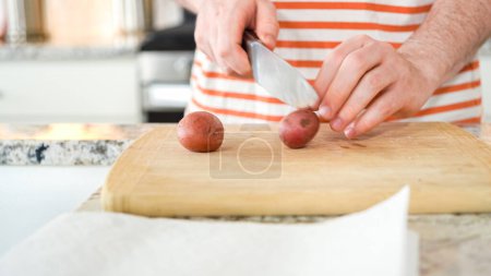 In the contemporary ambiance of a modern kitchen, a young man engages in dinner preparations. His current activity entails meticulously slicing small rainbow potatoes in half on a wooden cutting board