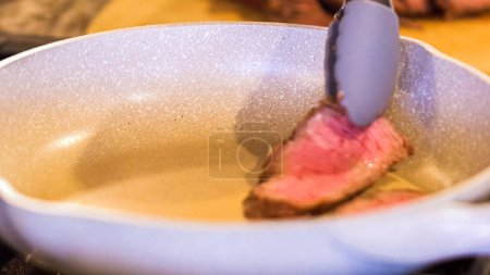 Téléchargez les photos : Pinces tournant de succulentes lanières de steak moyennement rares dans une poêle en céramique mouchetée. - en image libre de droit