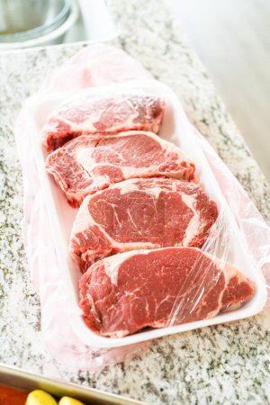 In einer eleganten, modernen weißen Küche wird ein hochwertiges Rib-Eye-Steak vorsichtig aus der Ladenverpackung entfernt. Das wunderschön marmorierte Steak, das bald gewürzt wird, verspricht eine bevorstehende kulinarische