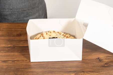 Foto de Colocar cuidadosamente el pastel de pan de jengibre, adornado con glaseado de caramelo salado y aspersiones de pan de jengibre, en una caja de pastel de paquete de papel blanco con una ventana transparente para regalar. - Imagen libre de derechos