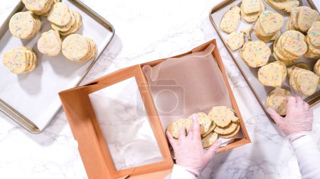 Téléchargez les photos : Pose plate. Avec précision, la femme arrange soigneusement les biscuits au sucre, remplis de saupoudres mélangées à la pâte, dans une boîte en papier brun rustique. - en image libre de droit