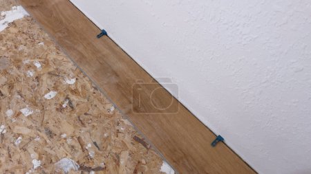 Ein einziger Vinyl-Dielenboden wird sorgfältig am Rand eines Raumes platziert, wobei Abstandshalter für eine präzise Passform an der strukturierten weißen Wand sorgen, was einen Schritt in der Transformation des Raumes markiert..