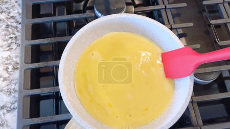 Hand holds a red spatula, stirring bright scrambled eggs in a non-stick frying pan, the blue flame of a gas stove underneath ensuring a warm, hearty meal.