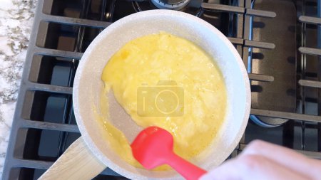 Hand holds a red spatula, stirring bright scrambled eggs in a non-stick frying pan, the blue flame of a gas stove underneath ensuring a warm, hearty meal.