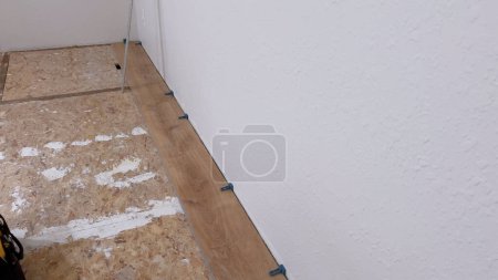 Téléchargez les photos : Un seul plancher en vinyle est soigneusement placé le long du bord d'une pièce, avec des entretoises assurant un ajustement précis contre le mur blanc texturé, marquant une étape dans la transformation de l'espace. - en image libre de droit