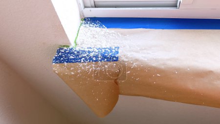 Meticulously masked edges and covered surfaces reveal the preliminary steps for painting a residential window sill. The contrast of blue tape against the sandy beige drop cloth illustrates a careful