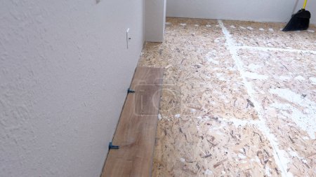 Un seul plancher en vinyle est soigneusement placé le long du bord d'une pièce, avec des entretoises assurant un ajustement précis contre le mur blanc texturé, marquant une étape dans la transformation de l'espace.