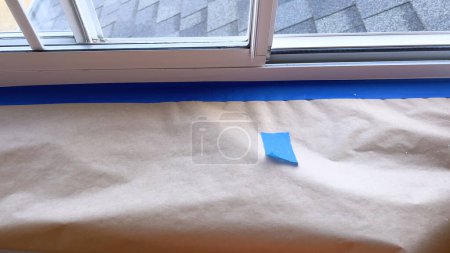 Téléchargez les photos : Des bords méticuleusement masqués et des surfaces couvertes révèlent les étapes préliminaires pour peindre un rebord de fenêtre résidentiel. Le contraste de la bande bleue contre le tissu de goutte beige sable illustre une attention particulière - en image libre de droit