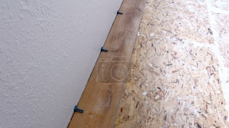 Un seul plancher en vinyle est soigneusement placé le long du bord d'une pièce, avec des entretoises assurant un ajustement précis contre le mur blanc texturé, marquant une étape dans la transformation de l'espace.