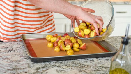 In a modern, white kitchen, a young man is engrossed in dinner preparations. His current endeavor includes arranging seasoned rainbow potatoes on a baking sheet, a meticulous step towards a