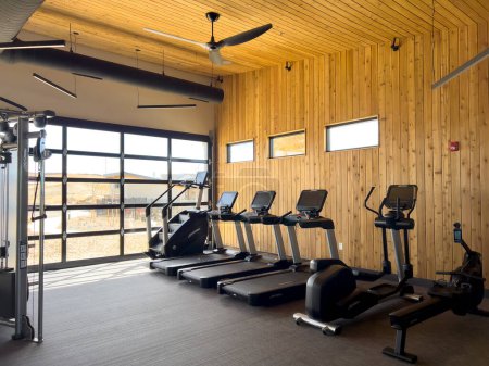 Foto de Denver, Colorado, USA-March 3, 2024-El espacioso gimnasio está equipado con una variedad de máquinas de ejercicios modernas, bañadas en luz natural desde grandes ventanales, con un cálido telón de fondo de madera. - Imagen libre de derechos