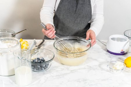 Téléchargez les photos : Intégrer les ingrédients dans un bol en verre, en remuant adepte avec un fouet pour confectionner un gâteau de bundt citron-myrtille alléchant. - en image libre de droit