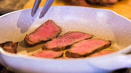 Téléchargez les photos : Pinces tournant de succulentes lanières de steak moyennement rares dans une poêle en céramique mouchetée. - en image libre de droit