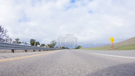An einem klaren Wintertag fährt ein Auto auf dem ikonischen Highway 101 in der Nähe von San Luis Obispo, Kalifornien. Die umliegende Landschaft ist bräunlich und gedämpft, mit sanften Hügeln und Küstenabschnitten