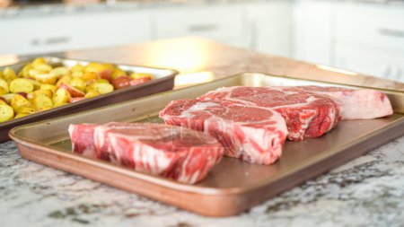 In the clean, minimalist layout of a modern white kitchen, dinner preparations are advancing smoothly. The ingredients, which include seasoned ribeye steaks and onion rings, are set out and ready to