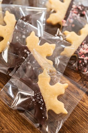 Embalaje cuidadoso galletas de recorte de Navidad, medio sumergidas en chocolate y presentadas en envoltura de celofán transparente, perfectas para regalos festivos.
