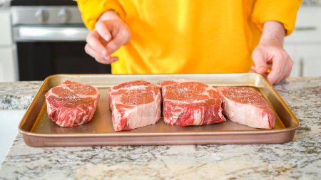 Photo for In the sophisticated layout of a modern kitchen, a young man is engrossed in dinner preparation. His current task involves seasoning large ribeye steaks, readying them for grilling, an essential step - Royalty Free Image
