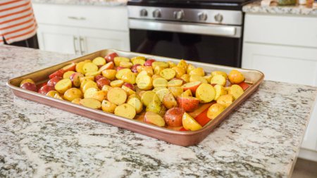 In a modern, white kitchen, a young man is engrossed in dinner preparations. His current endeavor includes arranging seasoned rainbow potatoes on a baking sheet, a meticulous step towards a
