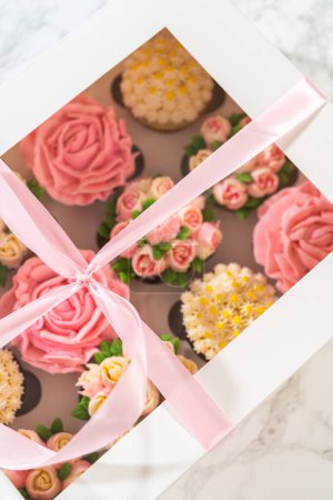 Téléchargez les photos : Enveloppé dans une boîte à cupcake en papier blanc immaculé, chaque cupcake gourmet est une œuvre d'art, ornée de fleurs de glaçage à la crème au beurre magnifiquement conçues pour ressembler à des roses et des tulipes vibrantes.. - en image libre de droit