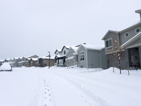Téléchargez les photos : Les rangées de maisons modernes sont recouvertes de neige, avec des pas qui tracent le seul chemin sur une rue étouffée par une lourde chute de neige sereine, la quiétude de la banlieue encapsulée dans une scène hivernale. - en image libre de droit