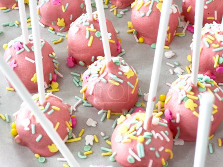 Arrayed neatly on parchment, these hand-dipped pink cake pops are a playful treat, adorned with a rainbow of sprinkles that add a festive touch to the sweet delights.