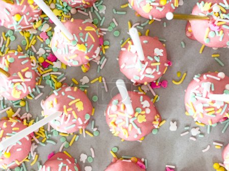 Fein säuberlich auf Pergament aufgereiht, sind diese von Hand eingetauchten rosa Cake Pops ein verspielter Leckerbissen, geschmückt mit einem Regenbogen aus Streusel, der den süßen Köstlichkeiten eine festliche Note verleiht..