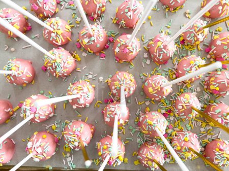 Arrayed neatly on parchment, these hand-dipped pink cake pops are a playful treat, adorned with a rainbow of sprinkles that add a festive touch to the sweet delights.
