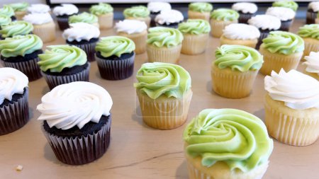 Filas de cupcakes recién horneados, mitad con chocolate negro y mitad con bases de vainilla, cada uno cubierto con glaseado blanco o verde maravillosamente remolinado, tientan los sentidos.