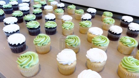 Rows of freshly baked cupcakes, half with dark chocolate and half with vanilla bases, each topped with beautifully swirled white or green frosting, tempt the senses.