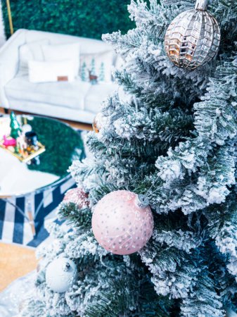 Im Vordergrund steht ein schneebedeckter, mit glitzernden Ornamenten geschmückter Weihnachtsbaum. Dahinter befindet sich ein stilvoller Wohnraum mit einer weißen Couch, einer grünen Wand und einem verspielten saisonalen Dekor.