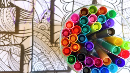 A kaleidoscopic collection of markers in a clear holder, poised above a detailed line drawing, invites a burst of artistic inspiration.