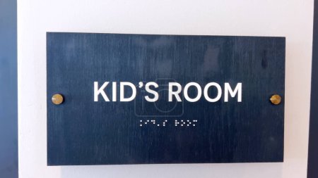 Eine dunkelblaue Gedenktafel an einer weißen Wand mit KIDS ROOM in weißen Großbuchstaben, die auf einen eigenen Kinderbereich hinweist.