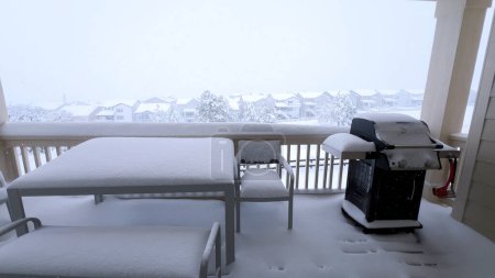 Thick layers of snow blanket a balcony with outdoor furniture, offering a serene view of a suburban landscape shrouded in winter embrace.