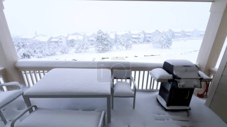 Foto de Capas gruesas de nieve cubren un balcón con muebles de exterior, ofreciendo una vista serena de un paisaje suburbano envuelto en abrazos de invierno. - Imagen libre de derechos