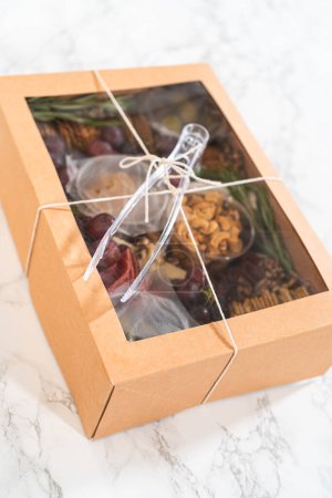Téléchargez les photos : Boîte de charcuterie avec viande tranchée, fromage, craquelins et raisins, le tout soigneusement emballé dans une boîte à cadeaux marron. - en image libre de droit