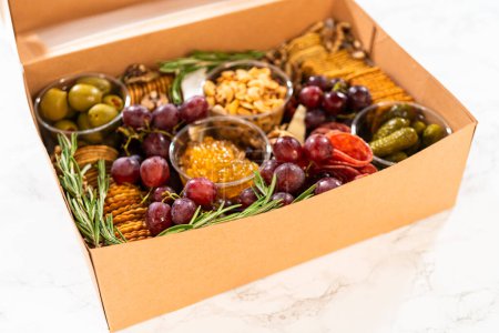 Téléchargez les photos : Boîte de charcuterie avec viande tranchée, fromage, craquelins et raisins, le tout soigneusement emballé dans une boîte à cadeaux marron. - en image libre de droit