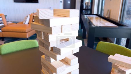 Ein unsicher gestapelter Turm aus Holzklötzen steht im Mittelpunkt und fordert die Spieler in einem Geschicklichkeits- und Gleichgewichtsspiel heraus. Der verschwommene Hintergrund deutet auf einen komfortablen Aufenthaltsraum hin, der darauf wartet,