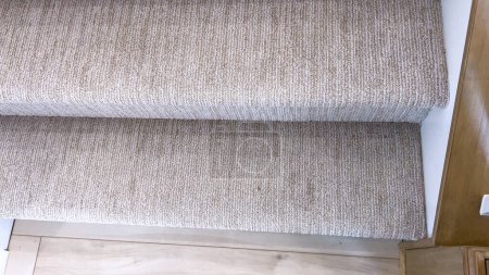 This image captures a detail of a home interior, focusing on the texture and pattern of beige carpeted stairs meeting a polished hardwood landing.