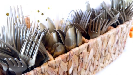Un primer plano de una cesta tejida llena de una variedad de tenedores grises y cucharas, preparado para un evento o buffet, que muestra un arreglo práctico y elegante utensilio.
