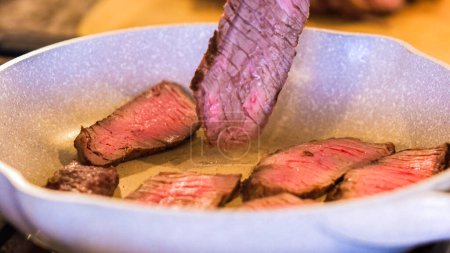 Photo for Tongs turning succulent medium-rare steak strips in a speckled ceramic skillet. - Royalty Free Image