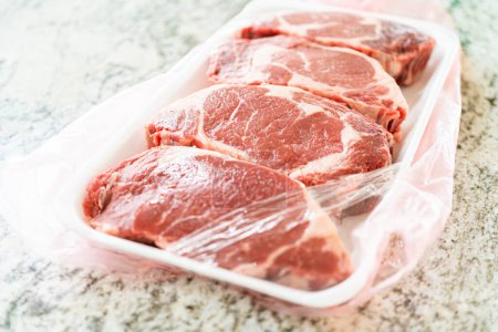In a sleek, modern white kitchen, a high-quality rib eye steak is carefully being removed from its store packaging. The beautifully marbled steak, soon to be seasoned, promises an upcoming culinary