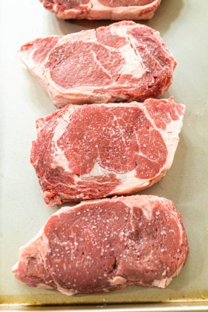 Situé dans une cuisine blanche moderne, un steak aux côtes assaisonné, vantant sa belle marbrure, se trouve prêt sur une plaque à pâtisserie. Il est préparé pour le barbecue à gaz extérieur, promettant une parfaite saisir sur le