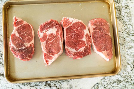 In einer modernen weißen Küche steht ein gewürztes Rib-Eye-Steak mit seiner schönen Marmorierung bereit auf einem Backblech. Es ist für den Outdoor-Gasgrill vorbereitet und verspricht einen perfekten Braten auf dem Grill