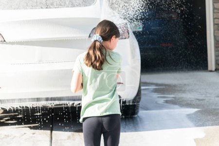 Una joven ayuda con entusiasmo a lavar el coche eléctrico de la familia en su entrada suburbana.