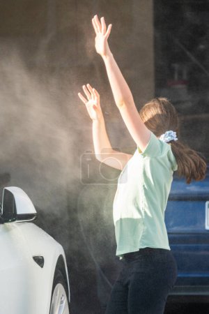 Una joven ayuda con entusiasmo a lavar el coche eléctrico de la familia en su entrada suburbana.